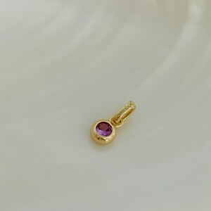 Puede incluir: Un colgante de tono dorado con una piedra preciosa rosa redonda y facetada engastada en un bisel liso y pulido. El colgante incluye una peque&ntilde;a anilla texturizada en la parte superior, marcada con el texto 925, indicando su base de plata.