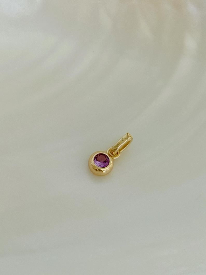 Puede incluir: Un colgante de tono dorado con una piedra preciosa rosa redonda y facetada engastada en un bisel liso y pulido. El colgante incluye una peque&ntilde;a anilla texturizada en la parte superior, marcada con el texto 925, indicando su base de plata.