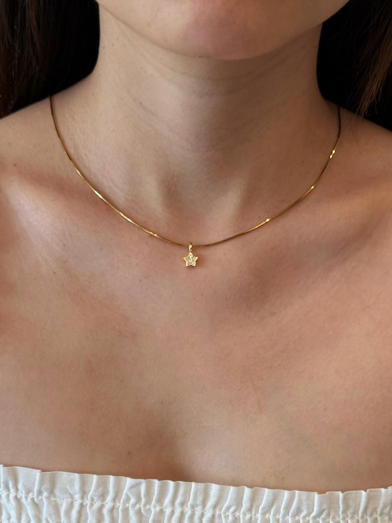 Op de afbeelding: Een dunne gouden ketting met een kleine, vijfhoekige sterhanger. De sterhanger is in het midden van de ketting geplaatst en bevat een enkele, kleine, heldere, fonkelende steen in het midden, rustend aan de basis van de nek.