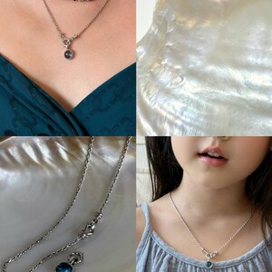 Pode incluir: Um colar de corrente de corda prateada com um pingente de pedra preciosa redonda de tom azul e uma arma&ccedil;&atilde;o de metal texturizado. O colar inclui um fecho de alavanca e &eacute; exibido sobreposto a um colar de contas multicolorido.