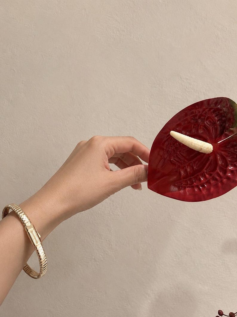 May include: A gold-toned metal bracelet with a flexible, ribbed link design and a rectangular clasp, worn on a wrist near a glossy, heart-shaped deep red anthurium flower featuring a prominent, textured, pale yellow spadix in the center.