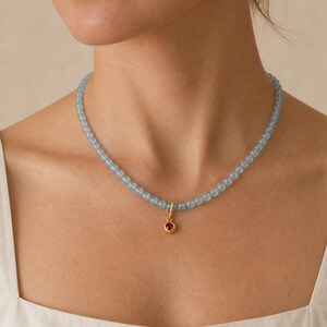 Puede incluir: Un collar de cuentas compuesto por peque&ntilde;as cuentas esf&eacute;ricas de color azul claro transl&uacute;cido ensartadas. Un colgante circular de tono dorado con una &uacute;nica piedra preciosa de color rojo vibrante engastada en el centro cuelga del medio.