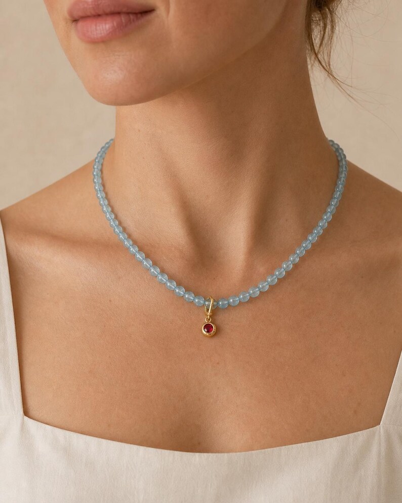 Puede incluir: Un collar de cuentas compuesto por peque&ntilde;as cuentas esf&eacute;ricas de color azul claro transl&uacute;cido ensartadas. Un colgante circular de tono dorado con una &uacute;nica piedra preciosa de color rojo vibrante engastada en el centro cuelga del medio.