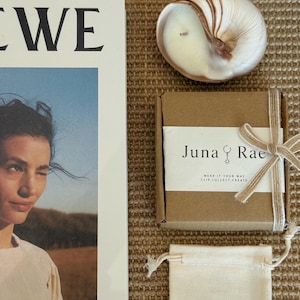 May include: A white magazine cover featuring a person, a shell-shaped candle, a brown cardboard box with a white label reading Juna Rae, and a small beige drawstring pouch. The label includes the text Wear It Your Way Clip.Collect.Create.