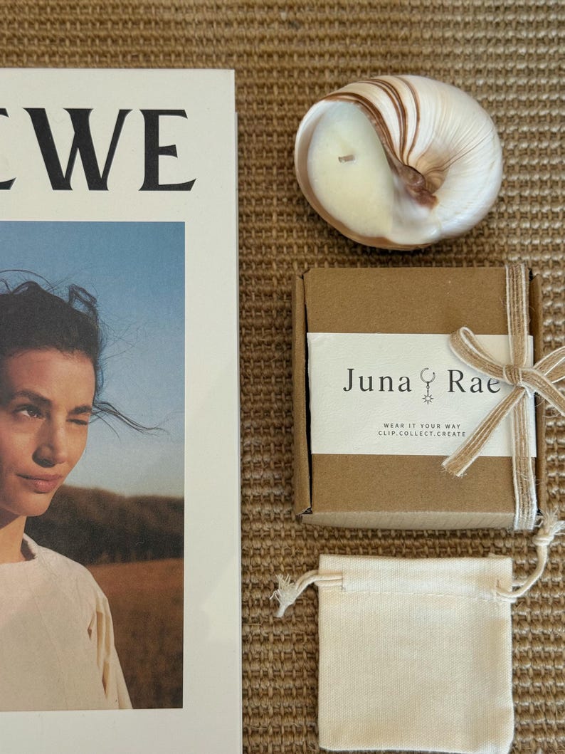 May include: A white magazine cover featuring a person, a shell-shaped candle, a brown cardboard box with a white label reading Juna Rae, and a small beige drawstring pouch. The label includes the text Wear It Your Way Clip.Collect.Create.
