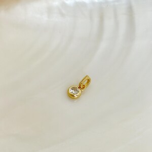 Puede incluir: Un peque&ntilde;o colgante de oro con una sola piedra preciosa redonda, transparente y facetada, engastada en un bisel de oro liso y pulido. El colgante incluye una peque&ntilde;a anilla de oro integrada en la parte superior para sujetarlo a una cadena.