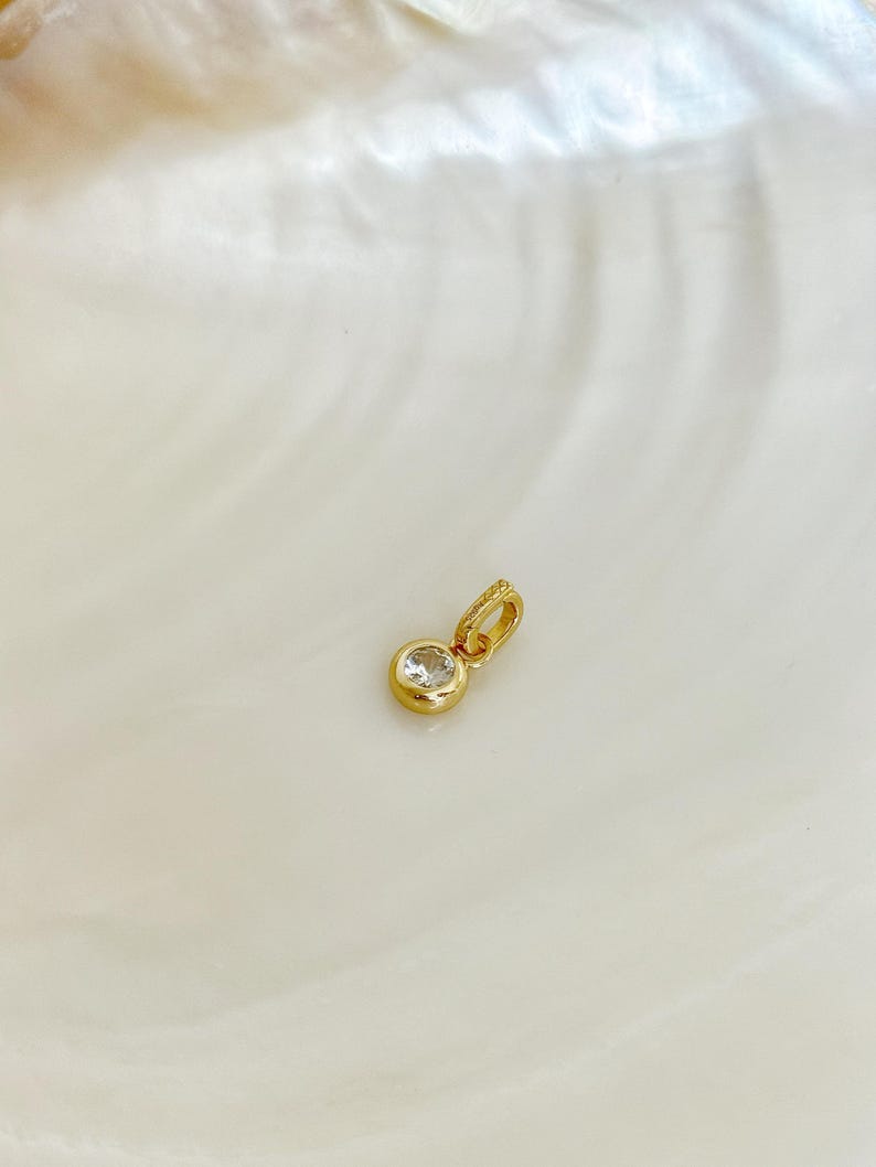 Puede incluir: Un peque&ntilde;o colgante de oro con una sola piedra preciosa redonda, transparente y facetada, engastada en un bisel de oro liso y pulido. El colgante incluye una peque&ntilde;a anilla de oro integrada en la parte superior para sujetarlo a una cadena.