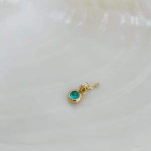 Puede incluir: Un peque&ntilde;o colgante de oro con una piedra preciosa redonda de color verde azulado engastada en bisel. El colgante tiene un acabado en oro pulido e incluye una peque&ntilde;a anilla integrada en la parte superior para sujetarlo a una cadena.