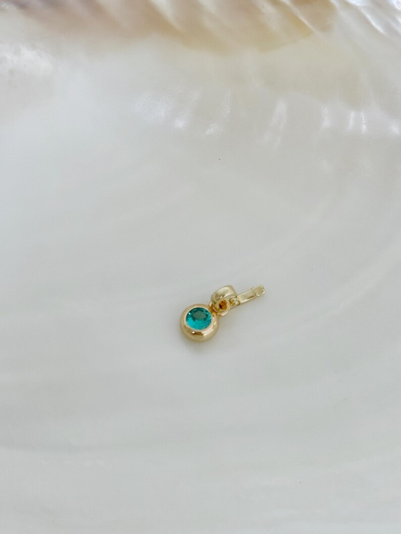 Puede incluir: Un peque&ntilde;o colgante de oro con una piedra preciosa redonda de color verde azulado engastada en bisel. El colgante tiene un acabado en oro pulido e incluye una peque&ntilde;a anilla integrada en la parte superior para sujetarlo a una cadena.