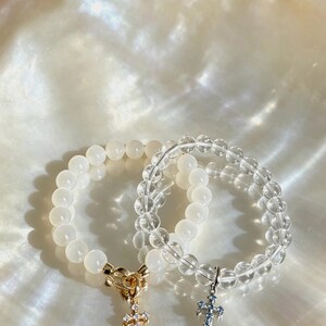 Puede incluir: Dos pulseras de cuentas redondas. Una pulsera tiene cuentas blancas opacas con un cierre de palanca dorado y un dije de cruz dorado con piedras claras. La otra pulsera tiene cuentas transparentes y un dije de cruz de tono plateado.
