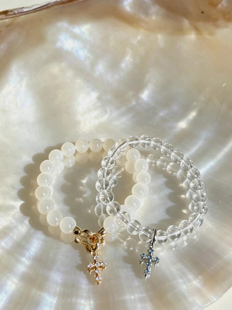 Puede incluir: Dos pulseras de cuentas redondas. Una pulsera tiene cuentas blancas opacas con un cierre de palanca dorado y un dije de cruz dorado con piedras claras. La otra pulsera tiene cuentas transparentes y un dije de cruz de tono plateado.