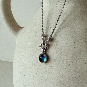 Pode incluir: Um colar de corrente de corda prateada com um pingente circular que apresenta uma pedra preciosa azul profunda no centro. O pingente &eacute; preso por um conector de metal texturizado e toda a pe&ccedil;a exibe um acabamento met&aacute;lico polido.