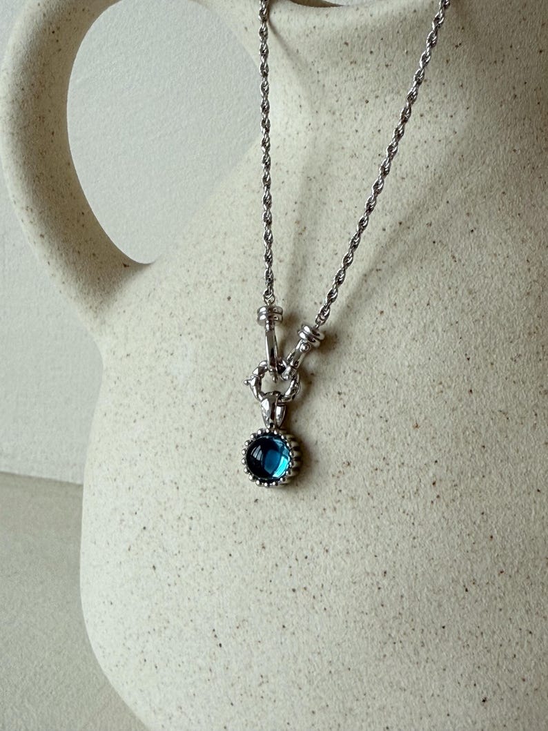 Pode incluir: Um colar de corrente de corda prateada com um pingente circular que apresenta uma pedra preciosa azul profunda no centro. O pingente &eacute; preso por um conector de metal texturizado e toda a pe&ccedil;a exibe um acabamento met&aacute;lico polido.