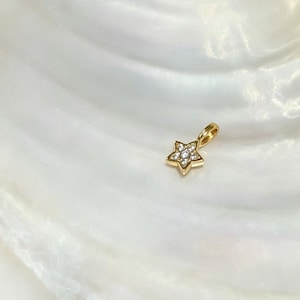 Op de afbeelding: Een kleine goudkleurige hanger in de vorm van een ster, voorzien van een centraal cluster van heldere, fonkelende steentjes. De ster heeft vijf punten en bevat een kleine, ge&iuml;ntegreerde gouden lus aan de bovenzijde voor bevestiging aan een ketting.
