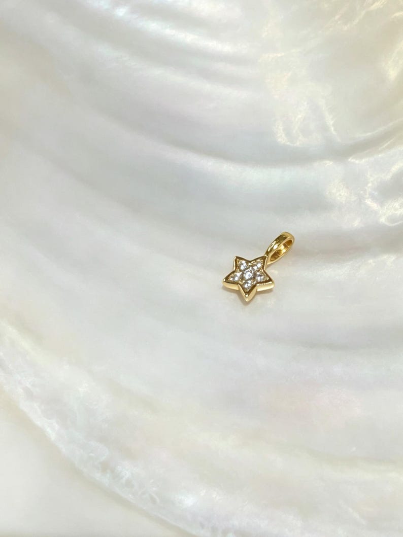 Op de afbeelding: Een kleine goudkleurige hanger in de vorm van een ster, voorzien van een centraal cluster van heldere, fonkelende steentjes. De ster heeft vijf punten en bevat een kleine, ge&iuml;ntegreerde gouden lus aan de bovenzijde voor bevestiging aan een ketting.