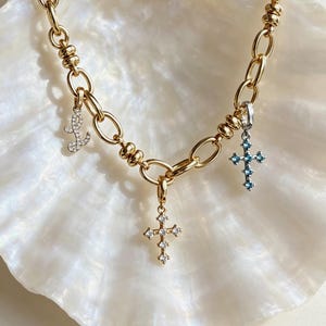 Puede incluir: Un collar de cadena en tono dorado con tres dijes colgantes: una letra L en cursiva incrustada con piedras transparentes, un dije de cruz con piedras transparentes y un dije de cruz con piedras azul claro. La cadena tiene eslabones ovalados.