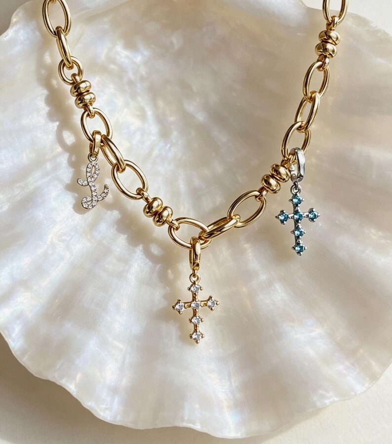 Puede incluir: Un collar de cadena en tono dorado con tres dijes colgantes: una letra L en cursiva incrustada con piedras transparentes, un dije de cruz con piedras transparentes y un dije de cruz con piedras azul claro. La cadena tiene eslabones ovalados.