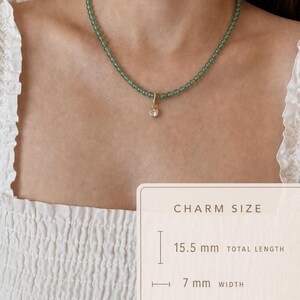 Puede incluir: Un collar con peque&ntilde;as cuentas redondas de color verde transl&uacute;cido y un dije dorado con una piedra redonda transparente. Una superposici&oacute;n indica que el dije mide 15,5 mm de largo total y 7 mm de ancho, incluyendo el cierre.