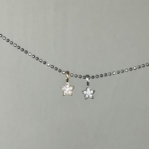 Op de afbeelding: Een zilverkleurige ketting met balletjes en twee kleine stervormige hangers. De ene hanger is goudkleurig en de andere is zilverkleurig, beide zijn bezet met kleine, heldere, fonkelende steentjes in het midden van elke ster.