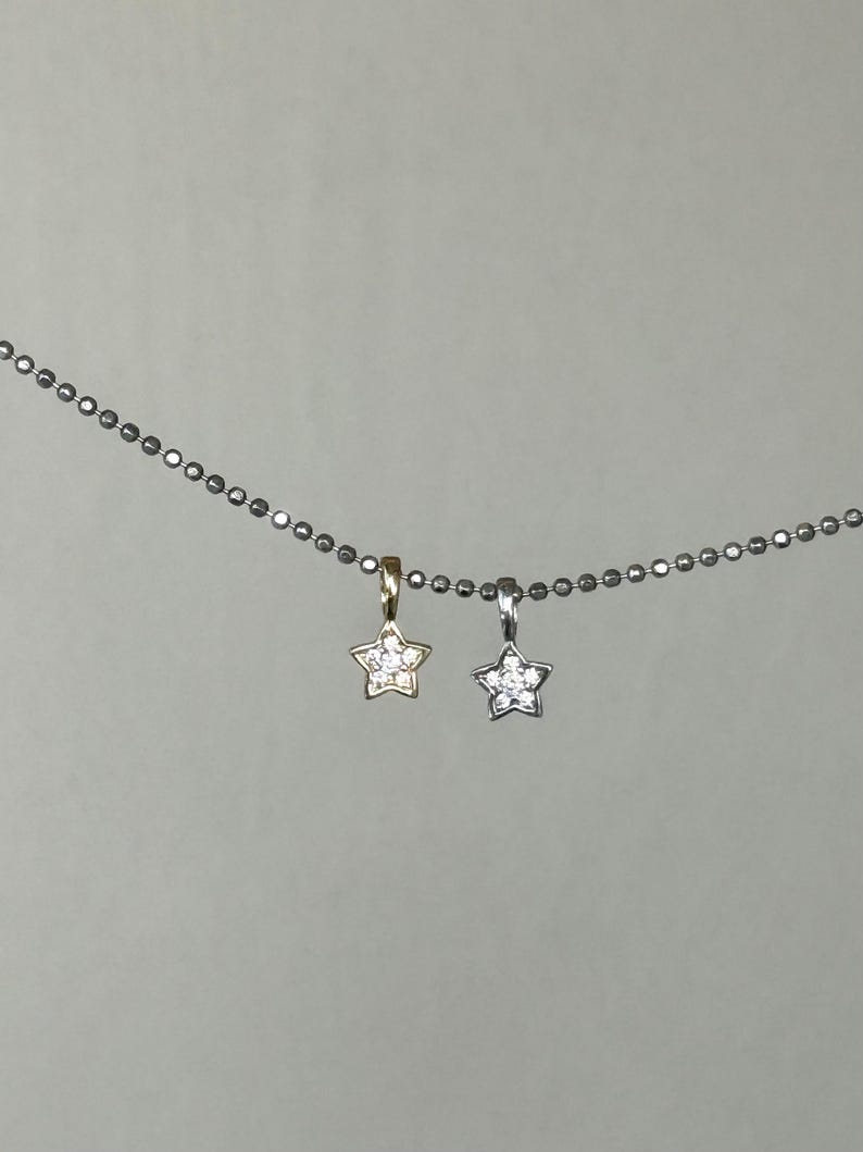 Op de afbeelding: Een zilverkleurige ketting met balletjes en twee kleine stervormige hangers. De ene hanger is goudkleurig en de andere is zilverkleurig, beide zijn bezet met kleine, heldere, fonkelende steentjes in het midden van elke ster.