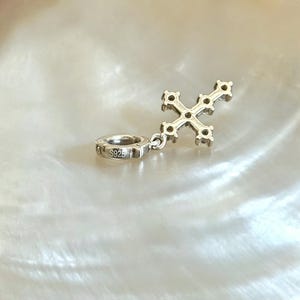 Puede incluir: Un dije de cruz de metal plateado con peque&ntilde;os engastes circulares a lo largo de sus brazos. El dije incluye un conector circular estampado con el texto S925, lo que indica una composici&oacute;n de plata esterlina. El dise&ntilde;o es met&aacute;lico.