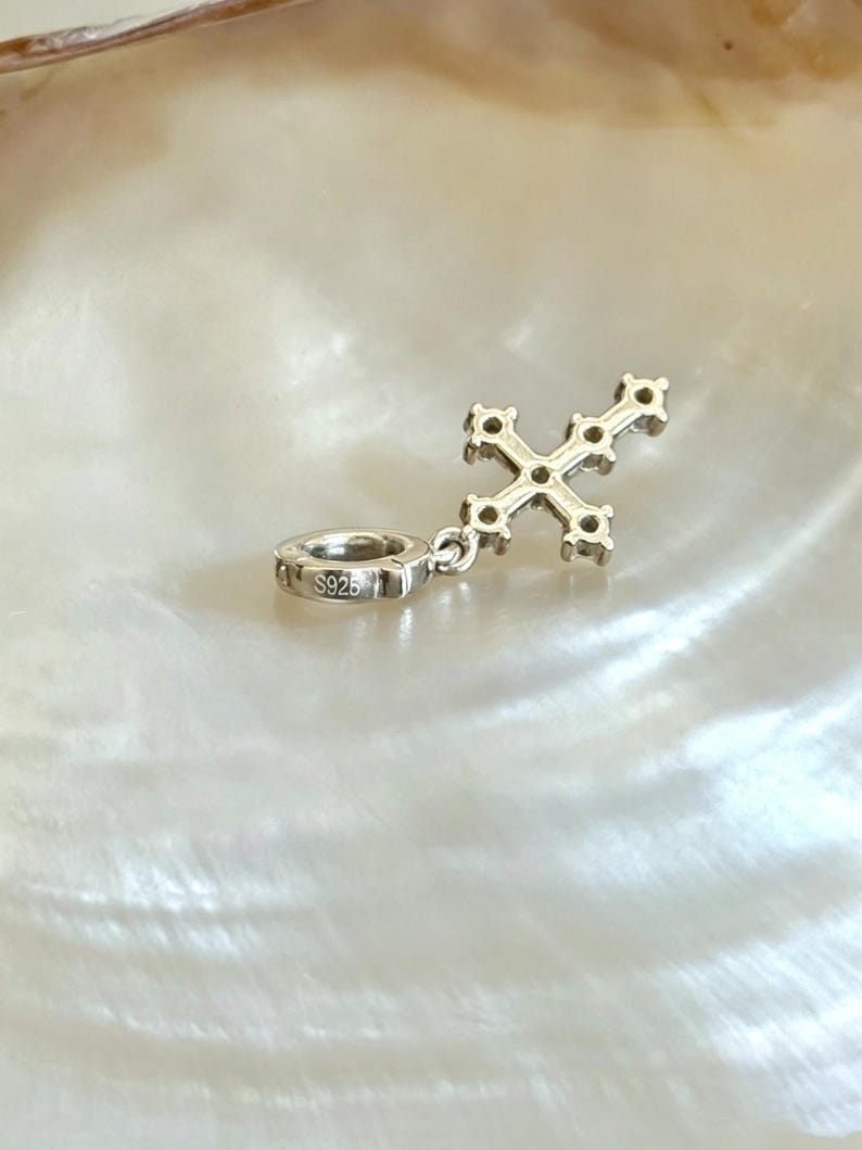 Puede incluir: Un dije de cruz de metal plateado con peque&ntilde;os engastes circulares a lo largo de sus brazos. El dije incluye un conector circular estampado con el texto S925, lo que indica una composici&oacute;n de plata esterlina. El dise&ntilde;o es met&aacute;lico.