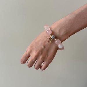 Puede incluir: Una pulsera de cuentas con esferas de cuarzo rosa p&aacute;lido, lisas y transl&uacute;cidas. La pulsera incluye un dije central de cruz en tono dorado adornado con peque&ntilde;as piedras claras y una cuenta esf&eacute;rica con una superficie plateada texturizada.