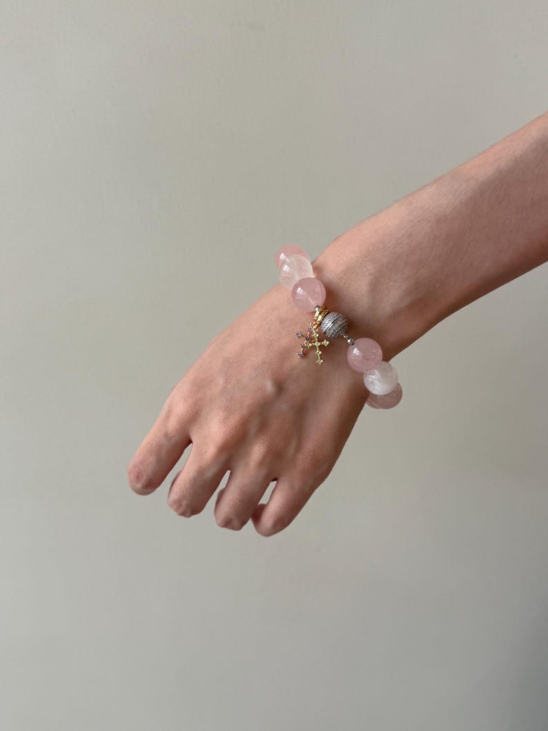 Puede incluir: Una pulsera de cuentas con esferas de cuarzo rosa p&aacute;lido, lisas y transl&uacute;cidas. La pulsera incluye un dije central de cruz en tono dorado adornado con peque&ntilde;as piedras claras y una cuenta esf&eacute;rica con una superficie plateada texturizada.