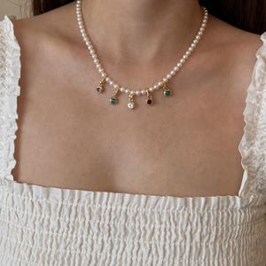 Puede incluir: Un collar que presenta una hilera de peque&ntilde;as perlas blancas y redondas. Suspendidos del centro de la hilera de perlas hay cinco peque&ntilde;os dijes dorados, cada uno engastado con una piedra preciosa redonda y facetada en colores morado, azul, transparente, rojo y verde.