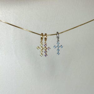 Puede incluir: Tres peque&ntilde;os colgantes de cruz colgados de una fina cadena dorada. Dos colgantes son de tono dorado con piedras preciosas de color verde claro y morado, mientras que el tercero es plateado con piedras azul claro en forma de cruz.