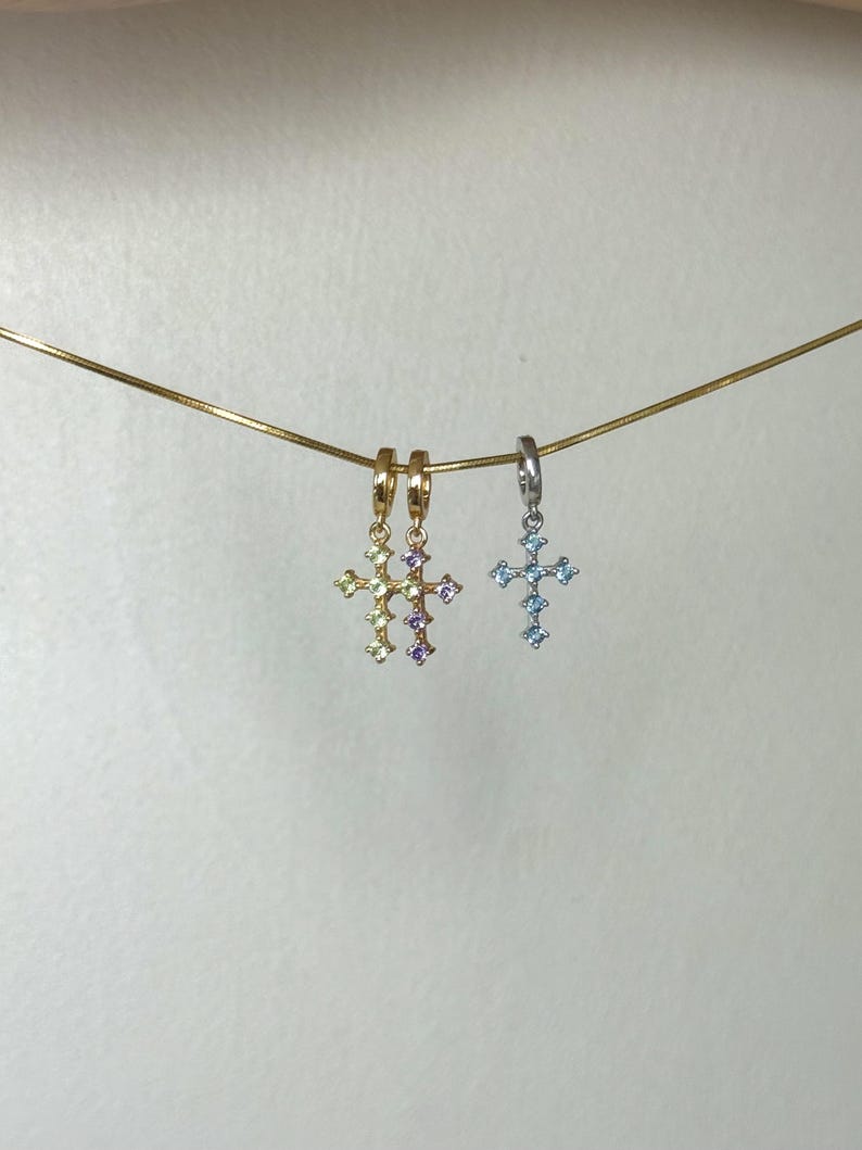 Puede incluir: Tres peque&ntilde;os colgantes de cruz colgados de una fina cadena dorada. Dos colgantes son de tono dorado con piedras preciosas de color verde claro y morado, mientras que el tercero es plateado con piedras azul claro en forma de cruz.