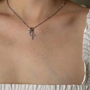 Op de afbeelding: Een zilverkleurige ketting met twee kleine bedels: een vijfpuntige ster ingelegd met heldere steentjes en een kruishanger die eveneens is bezet met heldere steentjes. De fijne metalen ketting rust op het gebied van het sleutelbeen.