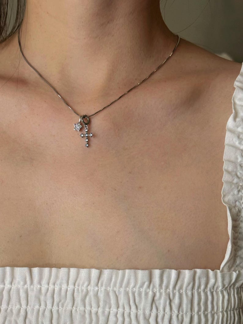 Op de afbeelding: Een zilverkleurige ketting met twee kleine bedels: een vijfpuntige ster ingelegd met heldere steentjes en een kruishanger die eveneens is bezet met heldere steentjes. De fijne metalen ketting rust op het gebied van het sleutelbeen.