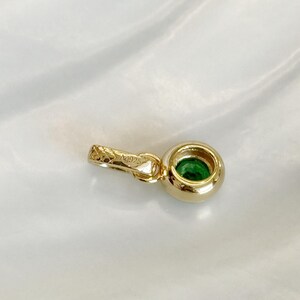 Puede incluir: Un colgante de tono dorado con una piedra preciosa verde redonda engastada en bisel. El colgante incluye una peque&ntilde;a anilla texturizada con el grabado Ag925 visible en el lateral. El dise&ntilde;o es sencillo y circular, destacando el contraste entre el metal dorado y la piedra verde vibrante.