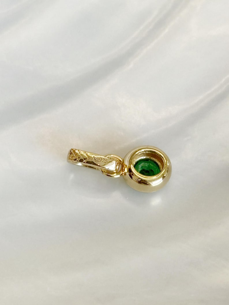 Puede incluir: Un colgante de tono dorado con una piedra preciosa verde redonda engastada en bisel. El colgante incluye una peque&ntilde;a anilla texturizada con el grabado Ag925 visible en el lateral. El dise&ntilde;o es sencillo y circular, destacando el contraste entre el metal dorado y la piedra verde vibrante.