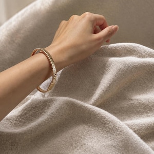 May include: A gold-toned bangle bracelet featuring a segmented, ribbed design with a polished finish. The bracelet has a circular shape and a uniform width, encircling the wrist with a series of repeating vertical metallic ridges.
