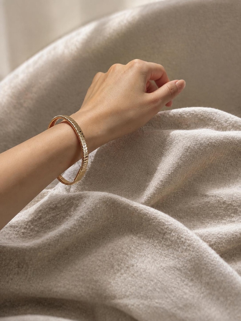May include: A gold-toned bangle bracelet featuring a segmented, ribbed design with a polished finish. The bracelet has a circular shape and a uniform width, encircling the wrist with a series of repeating vertical metallic ridges.