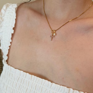 Puede incluir: Un collar de cadena en tono dorado con un peque&ntilde;o conjunto de dijes colgantes que consiste en una cruz delicada adornada con peque&ntilde;as piedras claras y una peque&ntilde;a estrella de cinco puntas con una piedra clara central, en un anillo dorado.