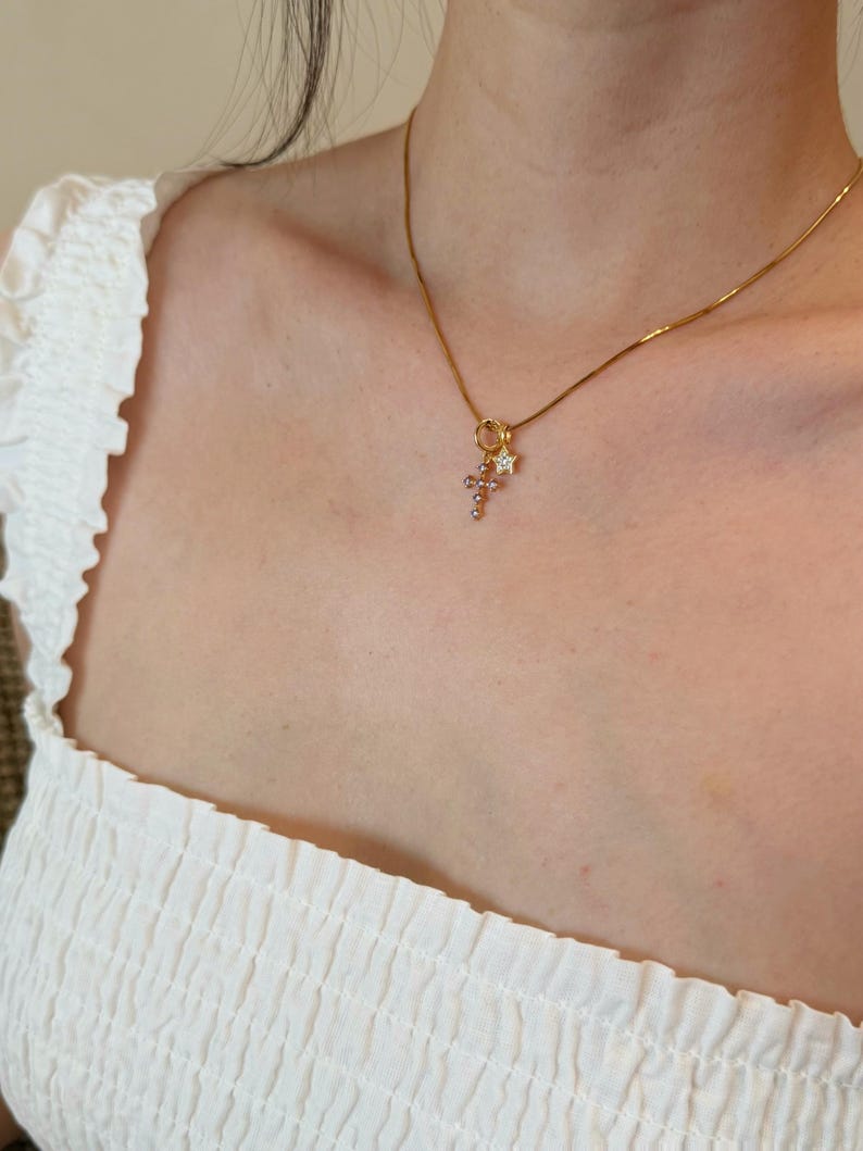 Puede incluir: Un collar de cadena en tono dorado con un peque&ntilde;o conjunto de dijes colgantes que consiste en una cruz delicada adornada con peque&ntilde;as piedras claras y una peque&ntilde;a estrella de cinco puntas con una piedra clara central, en un anillo dorado.