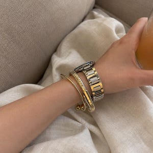 May include: A wrist adorned with three pieces of jewelry: a thin gold-tone bangle with spherical ends, a textured gold-tone cuff bracelet with a ribbed surface, and a two-tone silver and gold metal link watch with a round face and dark dial.