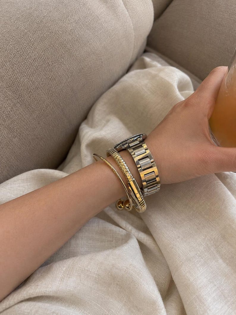 May include: A wrist adorned with three pieces of jewelry: a thin gold-tone bangle with spherical ends, a textured gold-tone cuff bracelet with a ribbed surface, and a two-tone silver and gold metal link watch with a round face and dark dial.