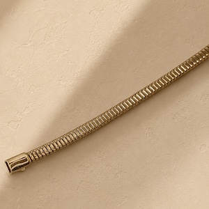 May include: A gold-toned snake chain necklace featuring a flexible, segmented metal construction with a smooth, polished rectangular clasp at the end. The chain has a uniform, cylindrical profile with a continuous series of interlocking links.