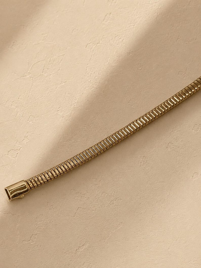 May include: A gold-toned snake chain necklace featuring a flexible, segmented metal construction with a smooth, polished rectangular clasp at the end. The chain has a uniform, cylindrical profile with a continuous series of interlocking links.