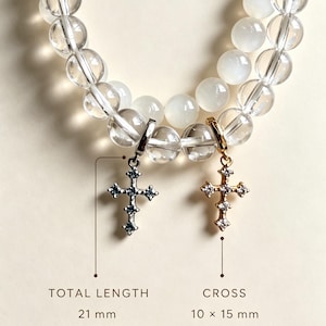 Puede incluir: Pulsera de cuentas esf&eacute;ricas transparentes y blancas con dos colgantes de cruz. Una cruz plateada y otra dorada, ambas con peque&ntilde;as piedras. El texto indica una longitud total de 21 mm incluyendo el cierre y un tama&ntilde;o de cruz de 10 x 15 mm.