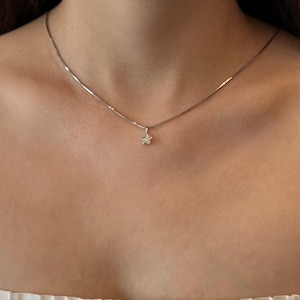 Op de afbeelding: Een delicate zilverkleurige ketting met een kleine, vijfpuntige sterhanger. De sterhanger is gecentreerd aan de dunne, gladde ketting en rust op het sleutelbeen, wat zorgt voor een subtiel en verfijnd metalen accessoire.