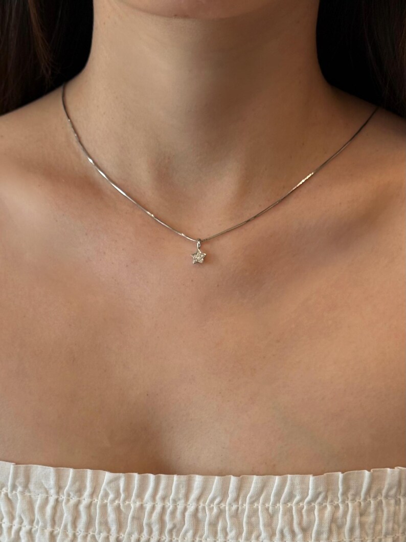Op de afbeelding: Een delicate zilverkleurige ketting met een kleine, vijfpuntige sterhanger. De sterhanger is gecentreerd aan de dunne, gladde ketting en rust op het sleutelbeen, wat zorgt voor een subtiel en verfijnd metalen accessoire.