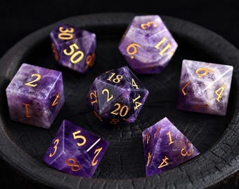 Puede incluir: Un juego de siete dados poliédricos tallados en piedra amatista morada con números grabados en color dorado. La colección incluye un d20, d12, d10, d8, d6, d4 y un dado de porcentaje, todos dispuestos en una bandeja circular de madera negra.