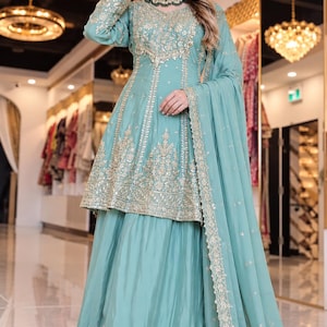 May include: A light teal peplum-style tunic and matching floor-length skirt set featuring intricate gold embroidery and sequin detailing. The outfit includes a coordinating sheer dupatta with a gold-embroidered border and a matching choker necklace.