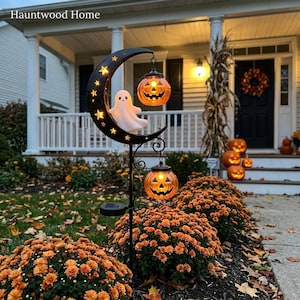May include: A black metal garden stake featuring a crescent moon silhouette with star cutouts, a white ghost figure, and two hanging orange jack-o'-lantern lights. The stake is positioned in a garden bed with orange mums and fallen leaves.