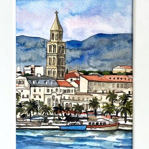 Może przedstawiać: Akwarela przedstawiająca nadmorskie miasto w białej ramie. Dzieło przedstawia krajobraz miejski z wysoką dzwonnicą, budynkami z czerwonymi dachami, palmami i łodziami na wodzie. Niebo jest niebieskie z nutą fioletu, a w tle góry.